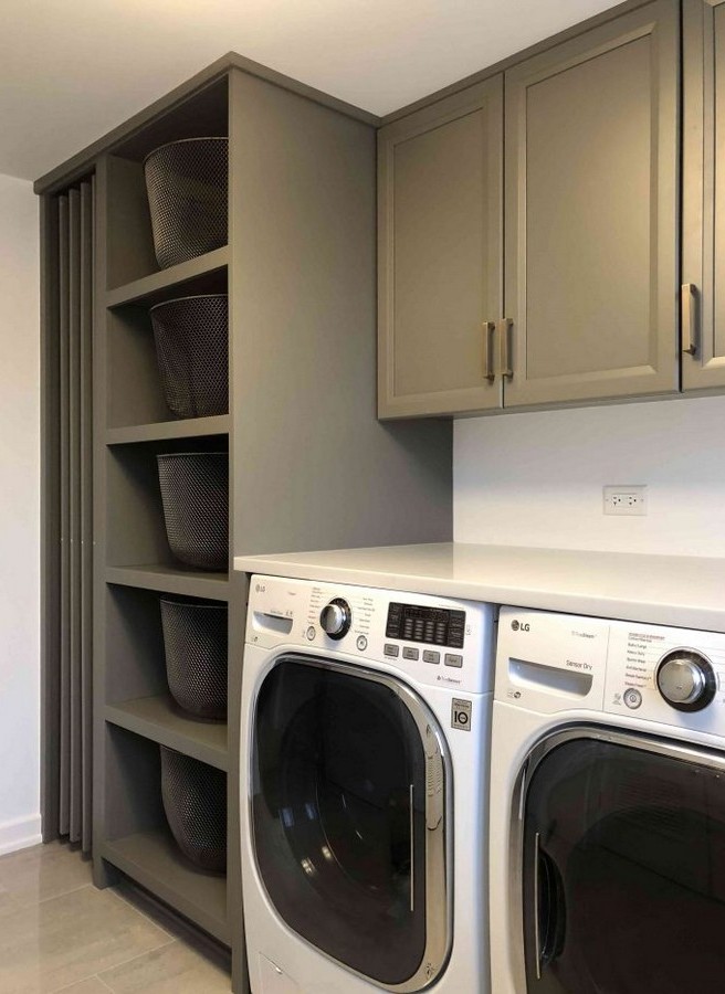 Laundry nook with front loading washer and dryer below a white countertop gray cabinetry and open shelving holding four gray baskets on the left