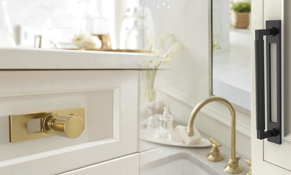 MedoraBackplates Woodworker Express Blog Close up of a white bathroom vanity with a gold faucet brass knob and marble countertop beside a glass door insight light filled room