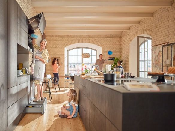 Open concept kitchen with a woman on a step stool reaching into upper cabinets while others cook and chat nearby