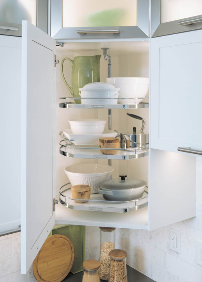 Open kitchen cabinet with three spiral rotating shelves holding bowls plates and cookware white and chrome with a green pitcher on top shelf
